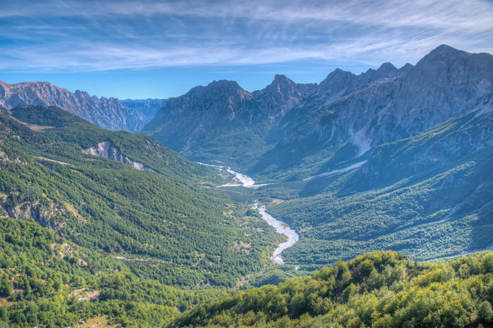 Aerial view of Valbona Valley, Albania, showing a winding river through forested mountains under a blue sky.