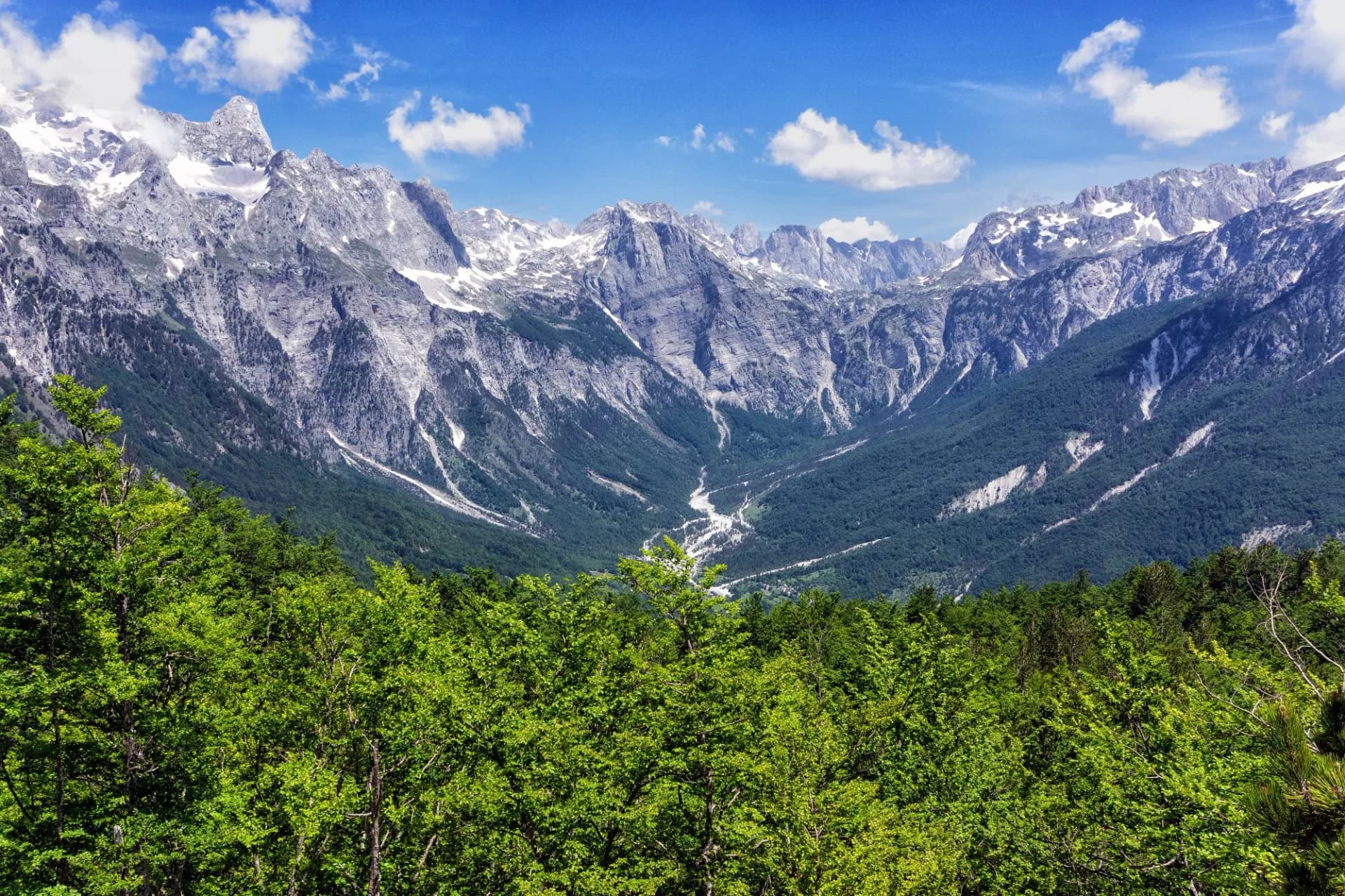 Majestic rocky Albanian Alps with snow patches towering over a green forested valley in Valbona Valley.