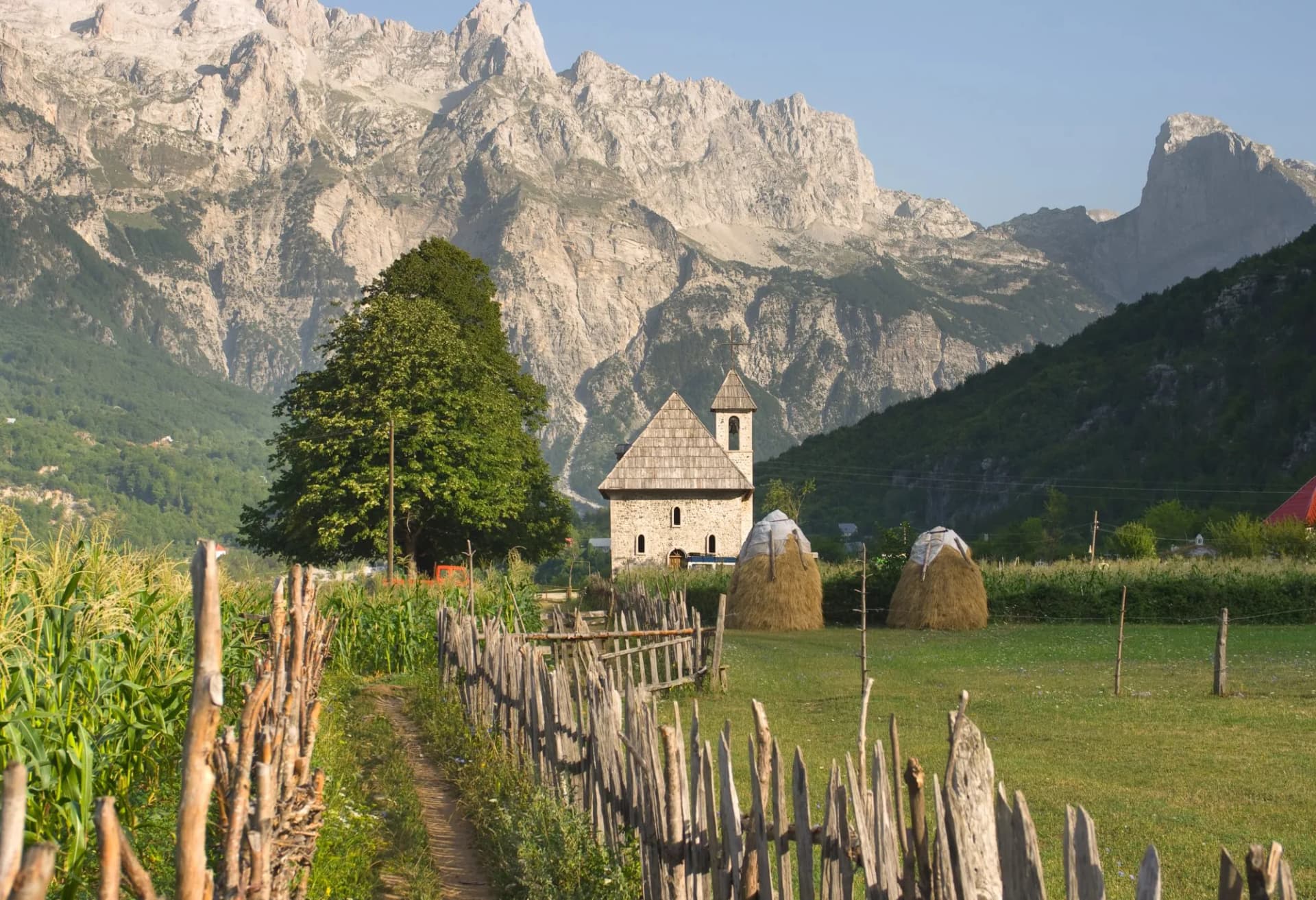 Stone church in Theth Valley, Albanian Alps, with cornfield and haystacks.