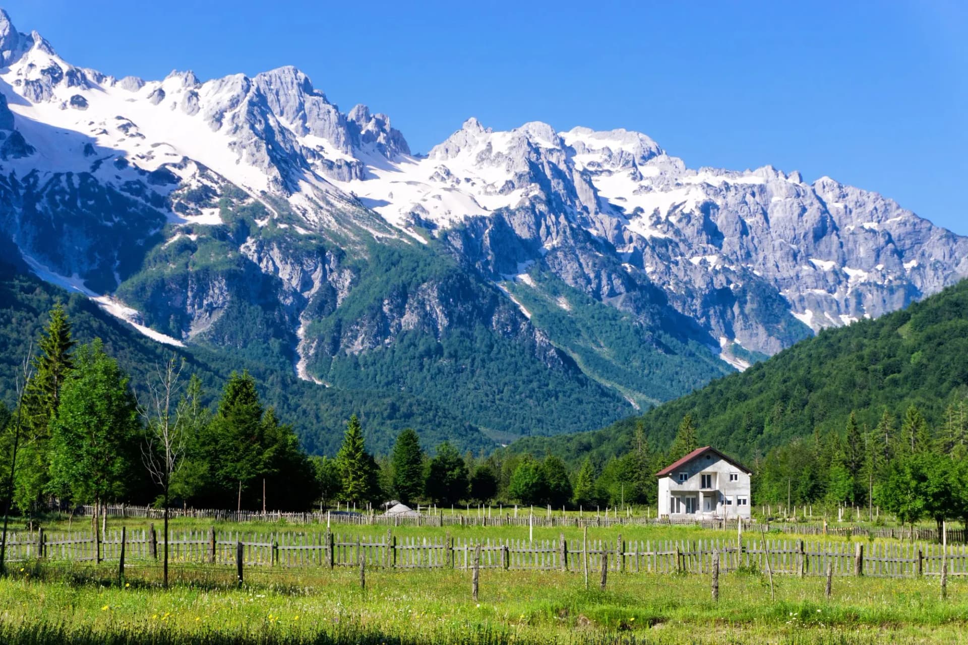 House in Valbona Village, Albania, with snow-capped mountains and green forest backdrop.