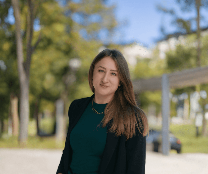 Woman in a dark green top and black blazer standing outdoors in a park setting.
