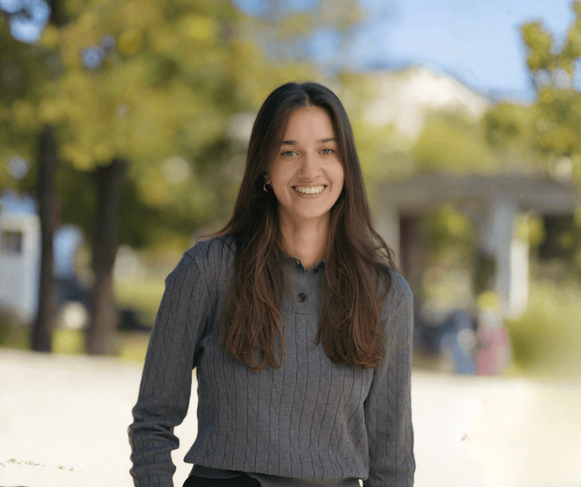 Young woman with long brown hair smiling outdoors in a park setting.