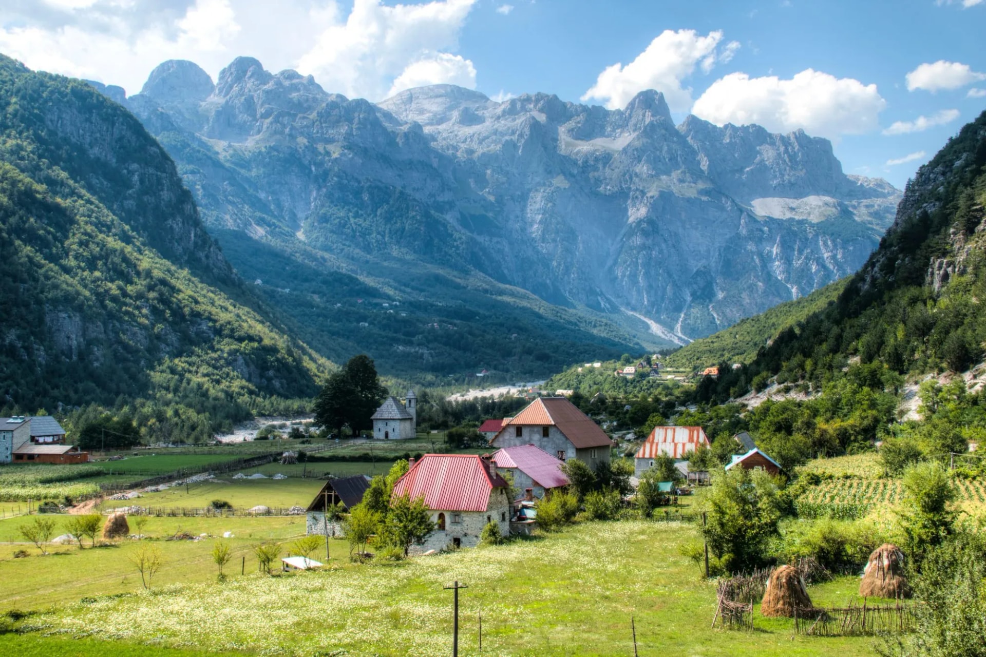 Mountain landscape in Theth with stone houses, green fields, and towering rocky peaks under a blue sky.