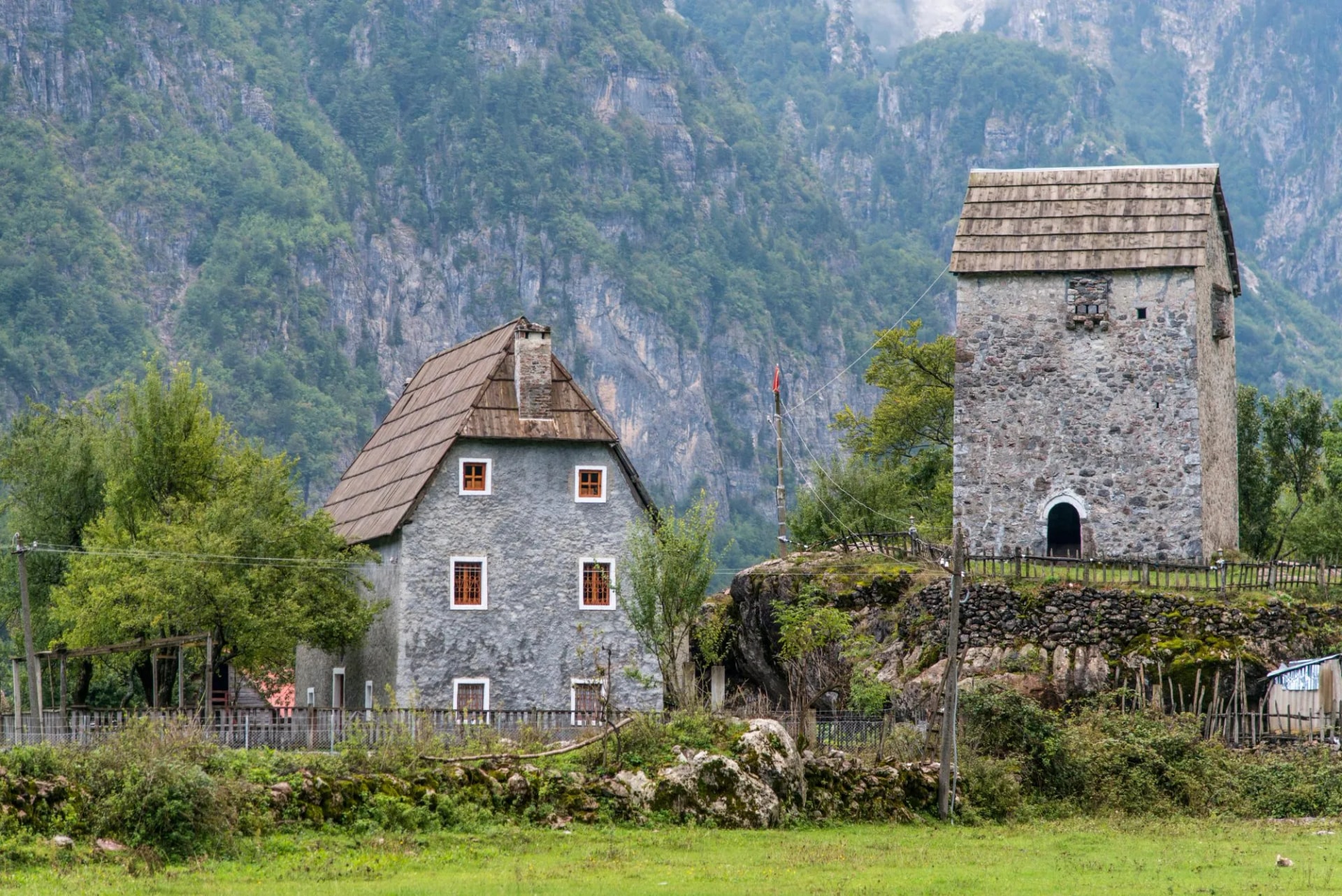 Stone tower and house in Theth National Park against steep, forested mountains.