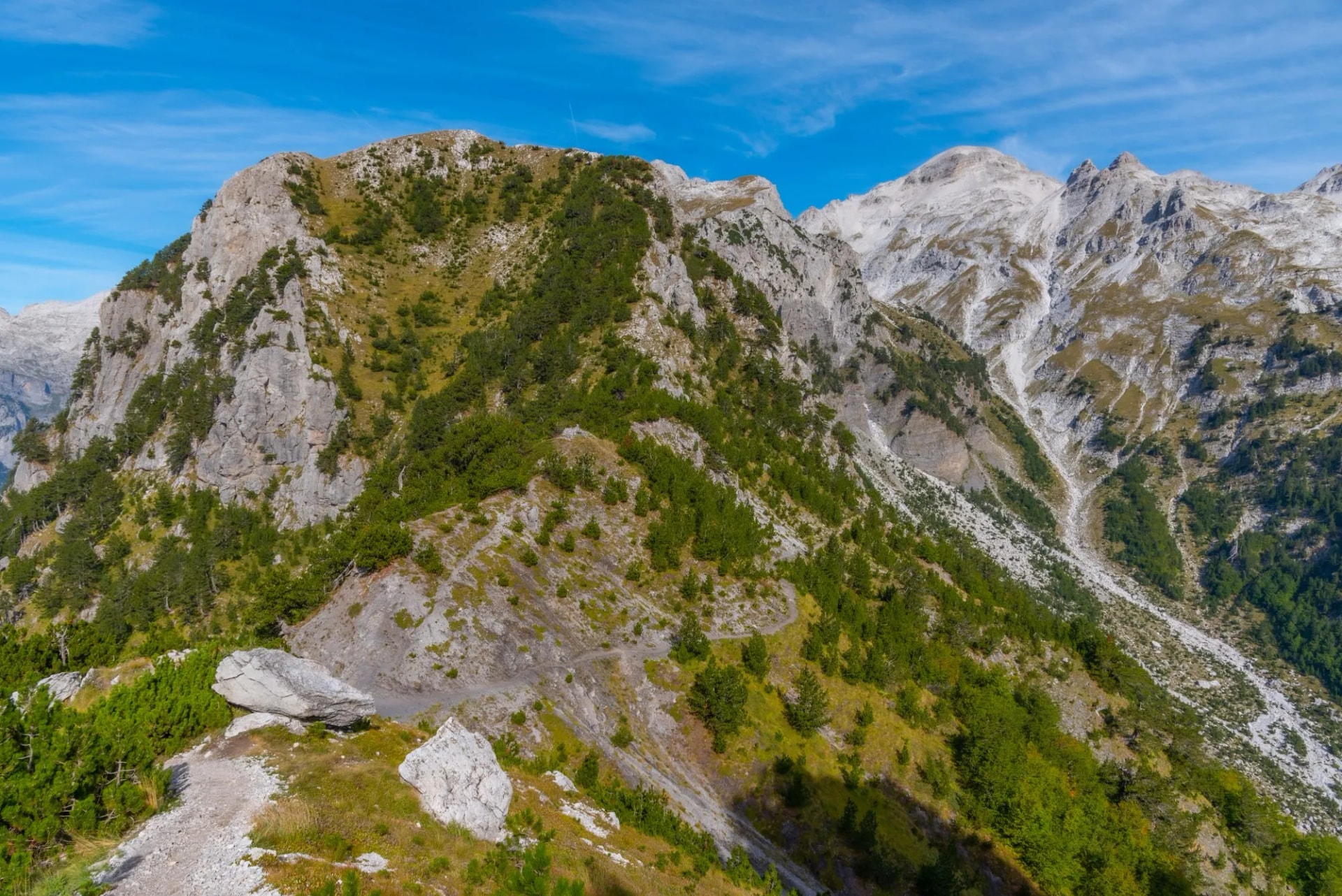 Hiking trail on rocky slope connecting Valbona and Theth valley mountains under blue sky.