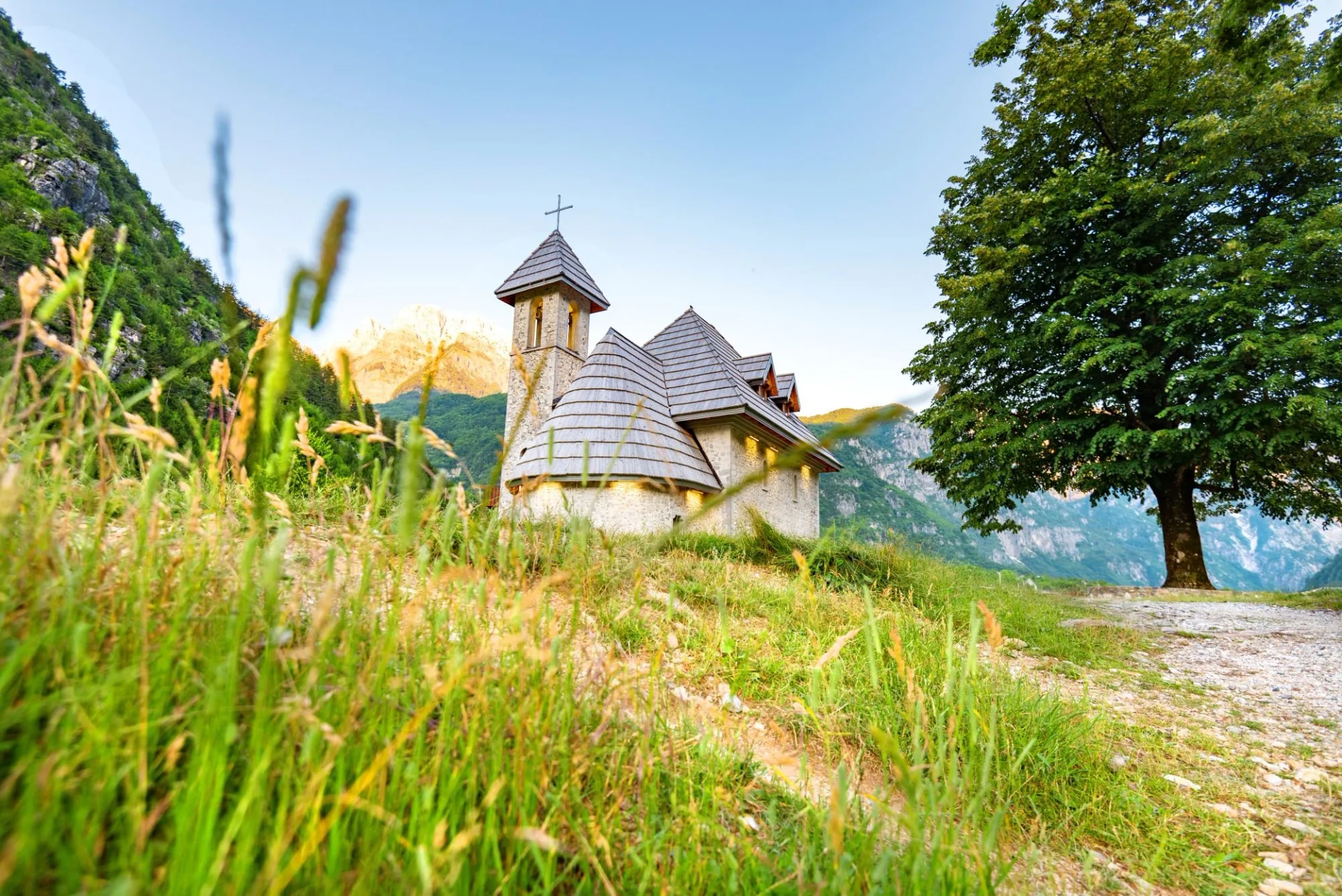 Stone church in Theth National Park on grassy hillside with mountains and large tree.