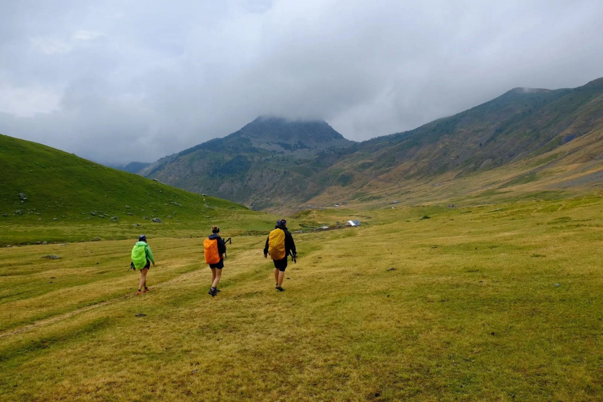 Hikers with backpacks walking through green valley toward mountains under cloudy sky