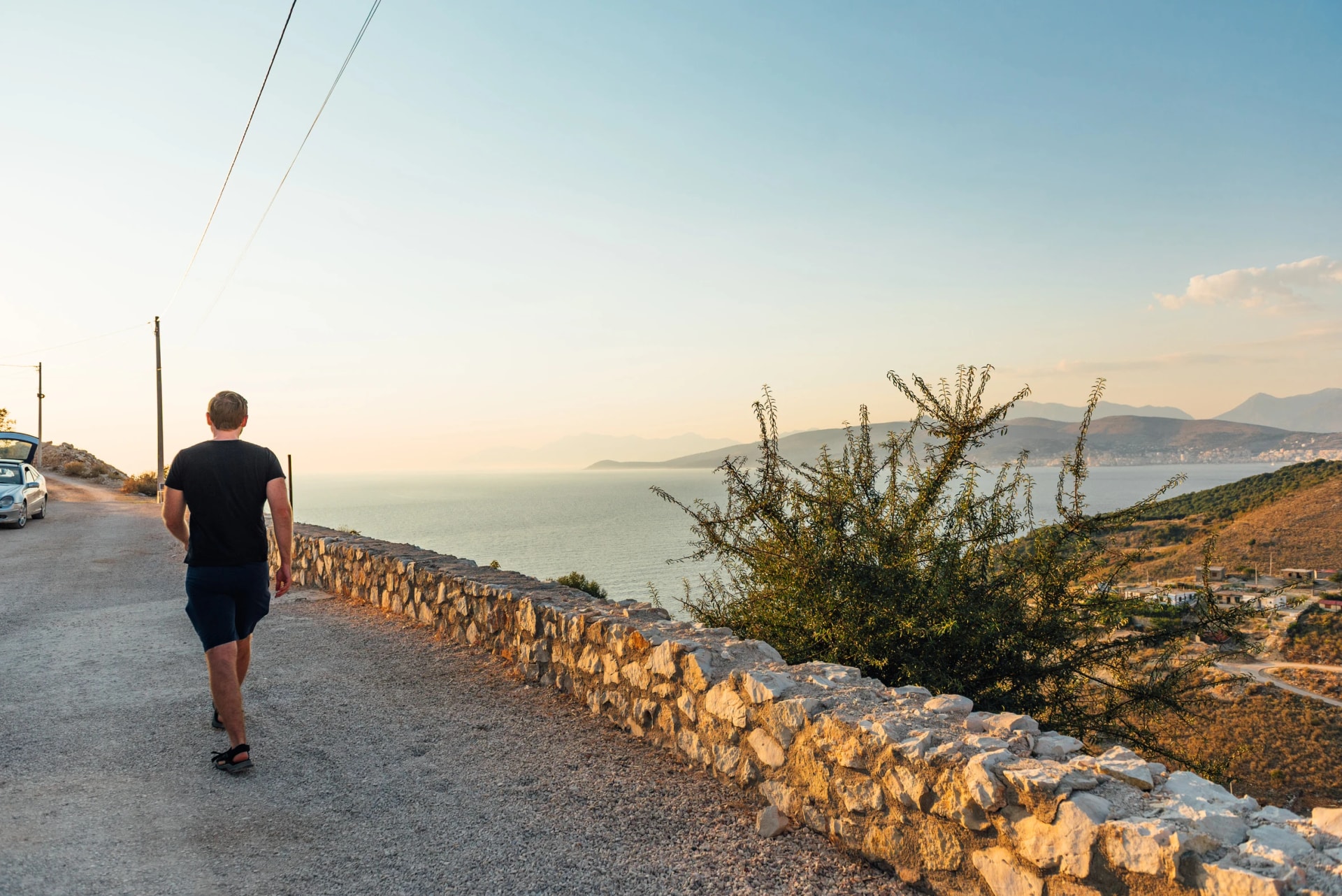 Traveler walking toward parked car on coastal road above Saranda Bay